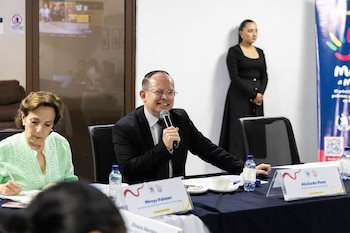 Un hombre con traje y corbata habla por micrófono sentado en una mesa con dos mujeres; una escribe, otra está de pie. Hay botellas de agua, tazas y carteles de nombres