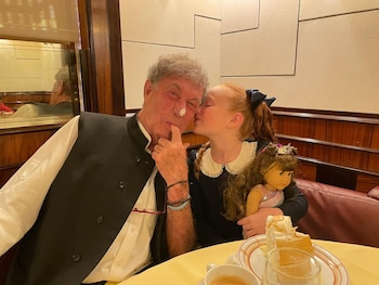 Alan Hassenfeld, con crema en la nariz, es besado en la mejilla por una niña pelirroja, Nicole, quien sostiene una muñeca. Ambos están en un restaurante