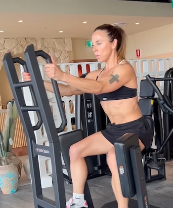 Una mujer atlética con top deportivo negro y shorts negros utiliza una máquina de gimnasio para entrenamiento de piernas. Tiene cabello recogido y un tatuaje en el brazo