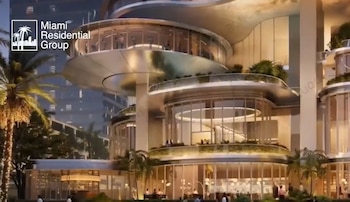Edificio moderno de múltiples niveles con fachadas de vidrio curvas, balcones, vegetación, palmeras y personas. En la esquina superior izquierda se ve el logo Miami Residential Group