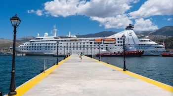 Vista de un gran barco blanco atracado en un muelle de hormigón con una franja amarilla y farolas negras. Se ven personas caminando por el muelle. Al fondo, montañas y cielo azul