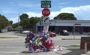 Globos de colores y peluches acomodados junto al poste de una señal de alto en una esquina de calle con edificios al fondo