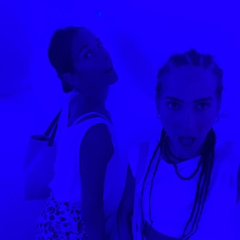 Juana Viale y Ambar de Benedictis, de pie, con expresiones sorprendidas, bajo una luz azul brillante que ilumina sus rostros y ropa clara