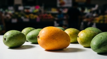 Un mango de color naranja y varios mangos verdes permanecen sobre una mesa blanca con un fondo oscuro desenfocado.