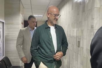 Un hombre calvo con gafas y camisa verde camina por un pasillo junto a un hombre con chaqueta beige