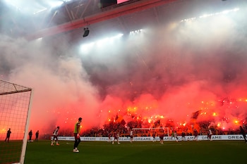 Jugadores en el campo de fútbol del estadio del Galatasaray, mientras la multitud en las gradas crea una nube de humo rojo intenso con bengalas y luces