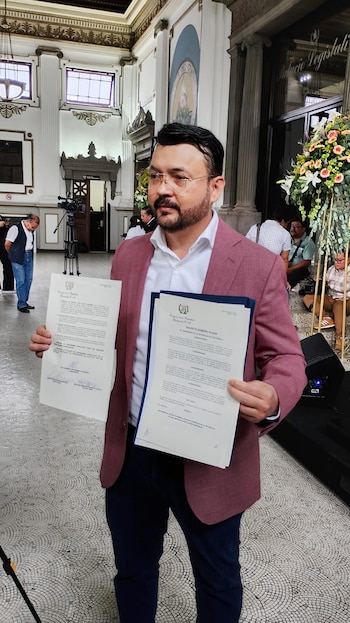 Hombre con barba, gafas y chaqueta rosa sostiene dos documentos. Detrás, el interior de un edificio con decoración floral, personas, una cámara y un cartel