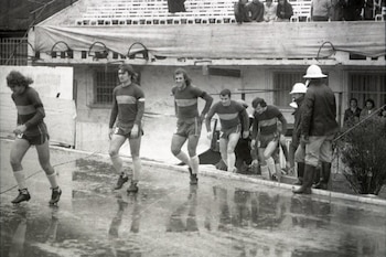Grupo de futbolistas con camiseta azul y amarilla, shorts y botines camina en suelo mojado. Hombres con casco y gorra los acompañan. Estructuras de estadio y público de fondo