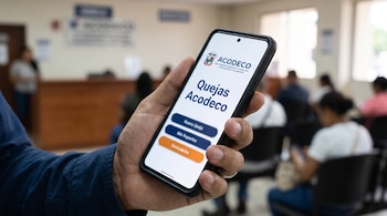 Una mano sostiene un teléfono inteligente con la pantalla de la aplicación Quejas Acodeco frente a personas sentadas en una sala de espera.