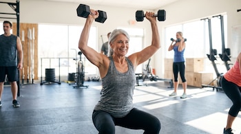 Mujer con cabello gris sonriendo en un gimnasio, haciendo sentadilla con mancuernas. Otros deportistas y equipamiento de gimnasio en el fondo.