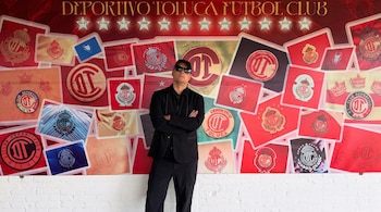 Hombre con gorra, gafas oscuras y ropa negra frente a un mural rojo cargado de escudos del Deportivo Toluca Fútbol Club.