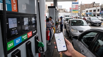 Una mano sostiene un teléfono inteligente frente a un surtidor de gas natural y gasolina con autos alineados al fondo.