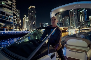 Hombre de mediana edad con camisa marrón oscuro pilotando una lancha rápida blanca por un canal urbano de noche, con rascacielos iluminados al fondo