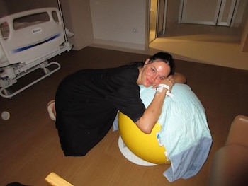 Mujer sonriente con bata negra y pulsera de hospital, apoyada en una pelota de parto amarilla en una habitación de hospital con una cama médica blanca