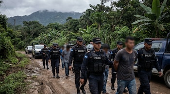 Grupo de policías y hombres con rostros desenfocados, algunos esposados, caminan por un camino de tierra embarrado. Varios vehículos policiales y vegetación densa al fondo.