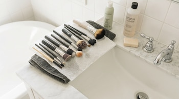 Doce brochas de maquillaje, dos botellas de limpiador de brochas, una barra de jabón, toallas de papel, una toalla gris, un grifo, un lavabo y una bañera.