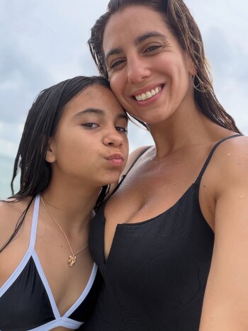 Una mujer con traje de baño negro sonríe junto a una joven que hace un gesto hacia la cámara en la playa