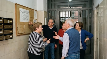 Cinco personas discuten frente a un tablón de anuncios con un cartel que dice "Se alquila espacio común" junto a buzones en el portal de un edificio.