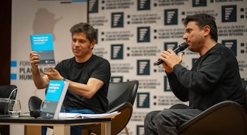 Dos hombres sentados en butacas negras ante una mesa baja; uno sostiene un libro azul y el otro habla por un micrófono frente a un panel