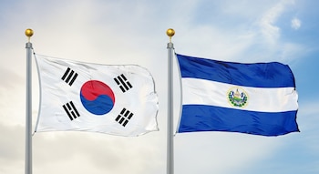 Banderas de Corea del Sur (blanca con círculo central rojo y azul) y El Salvador (azul y blanca con escudo) ondeando en mástiles contra el cielo.
