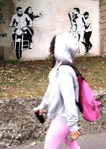 Una persona camina ante una pared blanca que contiene dos ilustraciones en blanco y negro con figuras humanas y una motocicleta
