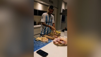 Hombre sonriente en una cocina moderna manipula piezas de pollo crudo sobre una tabla de cortar, con más pollo en bandejas y un teléfono celular en la mesada