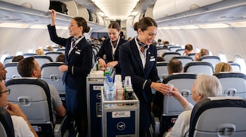 Tres tripulantes de cabina con uniforme azul atienden a pasajeros en el pasillo de un avión con un carrito de bebidas.