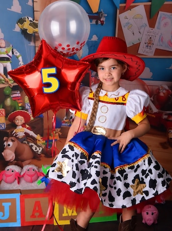 Una niña con un disfraz de vaquera sostiene globos frente a una decoración temática con personajes de dibujos animados y cubos con letras