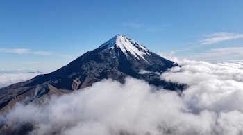 Una montaña con cumbre nevada sobresale por encima de capas de nubes blancas bajo un cielo azul.