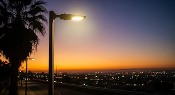 Una farola LED moderna brilla intensamente al atardecer con un cielo degradado de púrpura a naranja. La silueta de una palmera se ve a la izquierda y las luces borrosas de la ciudad de Los Ángeles en el fondo.
