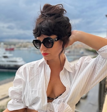 Primer plano de Griselda Siciliani usando gafas de sol negras, camisa blanca a rayas desabrochada y labial rojo, con un puerto al fondo