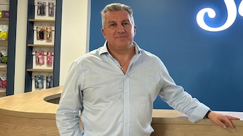 Hombre de mediana edad con camisa azul claro y pantalón oscuro posa junto a un mostrador de madera clara, con el logo iluminado de Saphirus en una pared azul de fondo