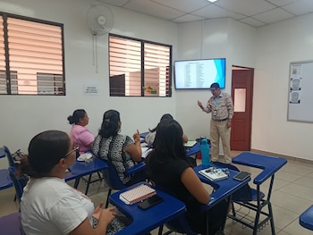 Personas sentadas en un aula gesticulan con las manos frente a una expositora, una pantalla y un pizarrón