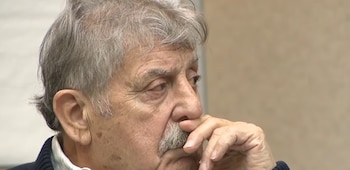 Un hombre mayor con cabello canoso y bigote sostiene su mano cerca de la cara mientras mira fijamente hacia un costado