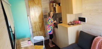 Mujer jubilada sonríe en la cocina de su mini casa. Viste camiseta colorida. La cocina tiene encimeras de madera clara, paredes de listones y azulejos decorativos