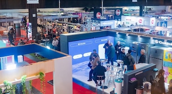 Gran salón de exposición con múltiples stands comerciales. Se observan personas, grandes pantallas, refrigeradores, lavadoras, y equipos diversos