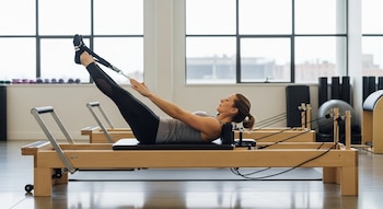 Mujer con ropa deportiva gris y negra realiza Pilates en una máquina reformer de madera clara, acostada de espaldas y levantando las piernas con correas en un estudio.