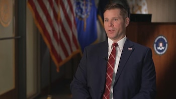 Primer plano del agente especial Doug Domin vistiendo traje oscuro y corbata roja, hablando con micrófono. De fondo, una bandera de EE. UU. y el logo del FBI