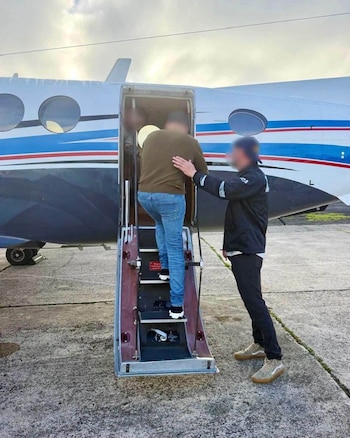Un hombre con el rostro difuminado sube la escalerilla de un avión pequeño junto a otra persona en una pista de aterrizaje