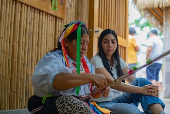 Dos mujeres sentadas, una indígena con atuendo tradicional teje con hilos de colores y la otra observa, frente a una pared de bambú
