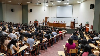 Vista panorámica de un auditorio universitario lleno de estudiantes sentados en pupitres de madera, observando un panel de oradores en un estrado frontal