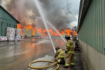 Tres bomberos, dos de ellos con mangueras, combaten un incendio industrial masivo con grandes llamas y denso humo oscuro que se eleva detrás de ellos en Maine