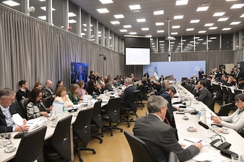 Personas sentadas en mesas dispuestas en fila en un salón de conferencias con micrófonos, documentos, pantallas y un logo institucional al fondo