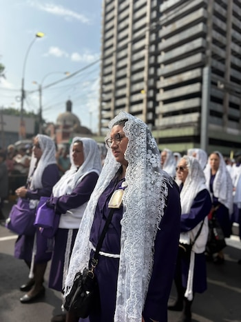 Mujeres vestidas con túnicas moradas y mantillas blancas de encaje permanecen de pie en una calle frente a edificios urbanos