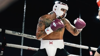 Un hombre con casco blanco, protector inguinal y guantes morados permanece en posición de guardia dentro de un cuadrilátero