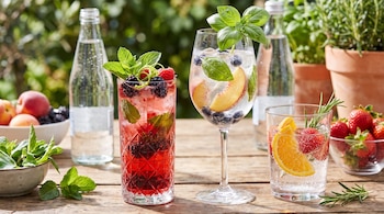 Tres vasos con bebidas de frutas y hierbas, dos botellas de agua mineral. Frutas varias, menta, albahaca, romero en tazones. Mesa de madera al aire libre.