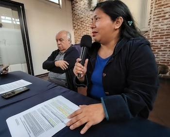 Mujer con cabello oscuro y chaqueta azul habla por micrófono. Hombre de cabello blanco y chaqueta oscura observa en segundo plano. Mesa con papeles y celular