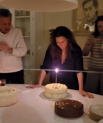 Una mujer se inclina hacia una tarta con una vela encendida en una mesa redonda con pasteles, junto a un hombre que aplaude