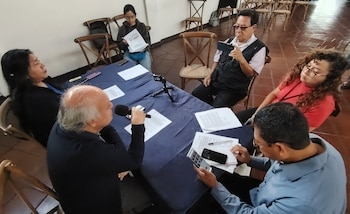 Seis personas sentadas alrededor de una mesa con mantel azul oscuro, papeles, dos micrófonos, tres teléfonos celulares, sillas de madera y piso de baldosas
