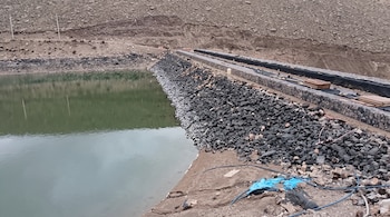 Un embalse de agua junto a un dique de rocas y gaviones en una zona árida con montañas al fondo.